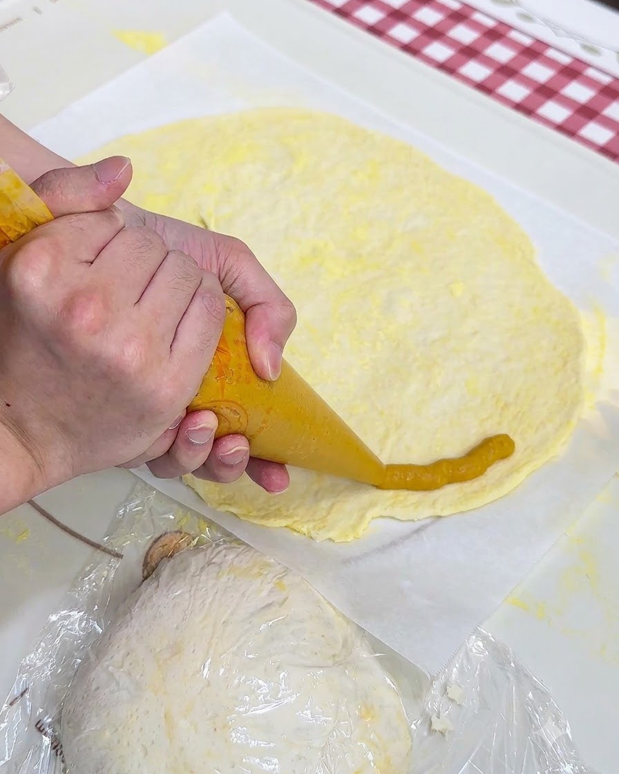 Mani che usano una sac à poche per formare un anello di purea di patate dolci attorno al bordo del disco di pasta per pizza su carta da forno.