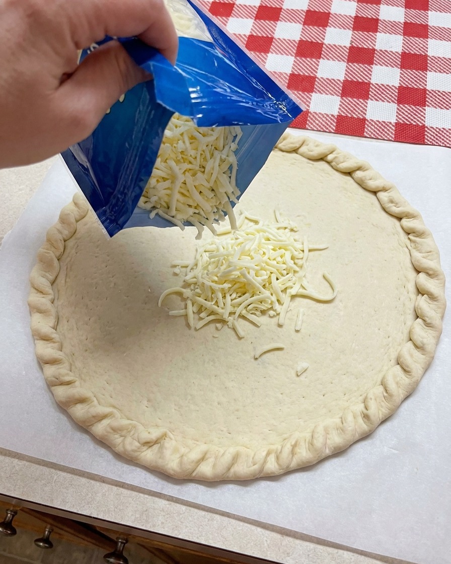 Mozzarella grattugiata versata da un sacchetto blu al centro di una base per pizza.