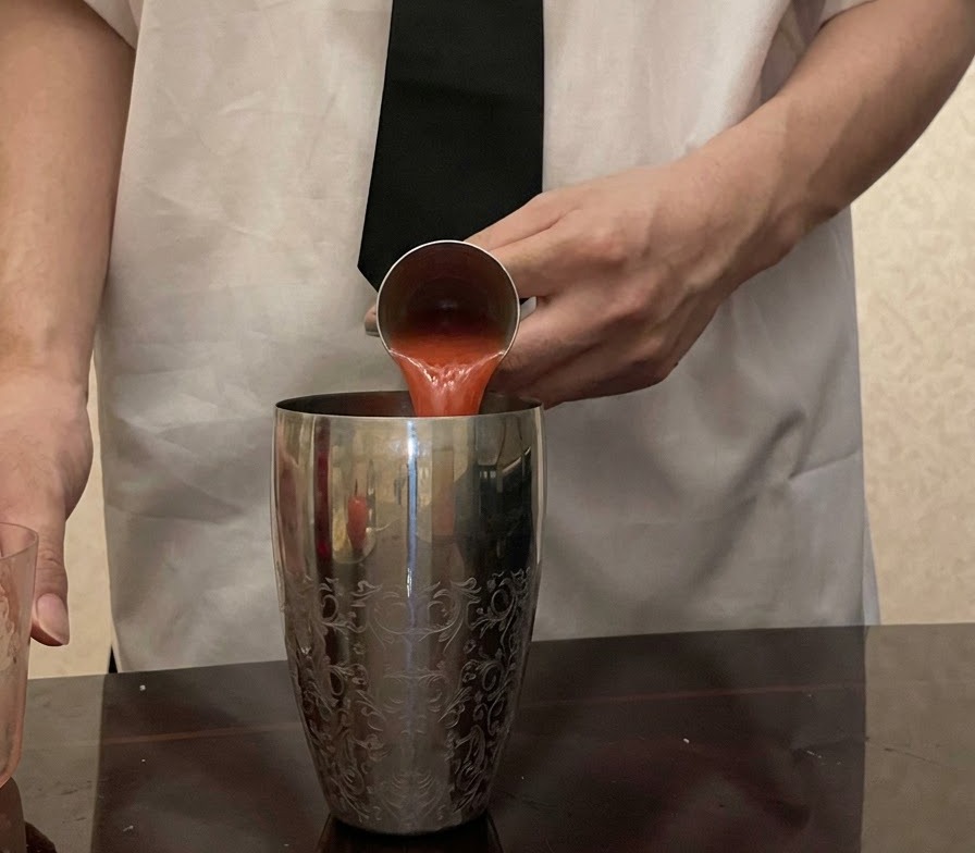 Succo di pomodoro rosso fresco versato da un misurino di metallo in uno shaker argentato fantasia.