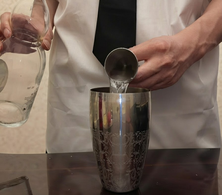 Tequila trasparente versata da una bottiglia di vetro in un misurino di metallo tenuto sopra uno shaker argentato.