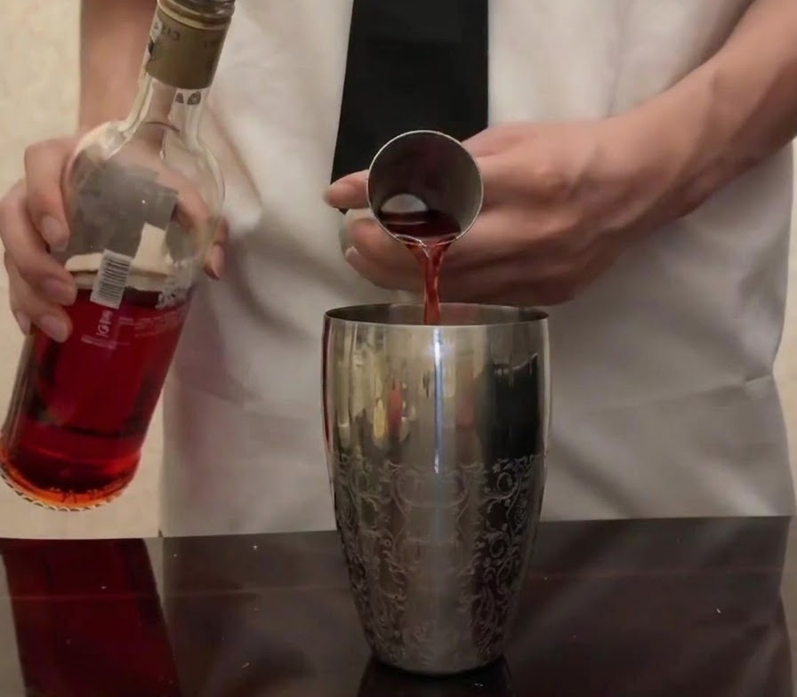 Campari rosso brillante versato dalla sua bottiglia in un misurino di metallo sopra uno shaker argentato.