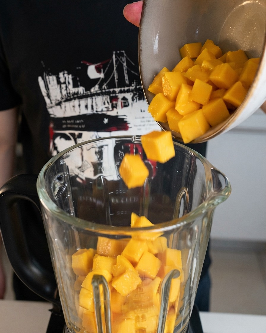 Cubetti di mango giallo fresco versati da una ciotola in un boccale di frullatore in vetro trasparente.