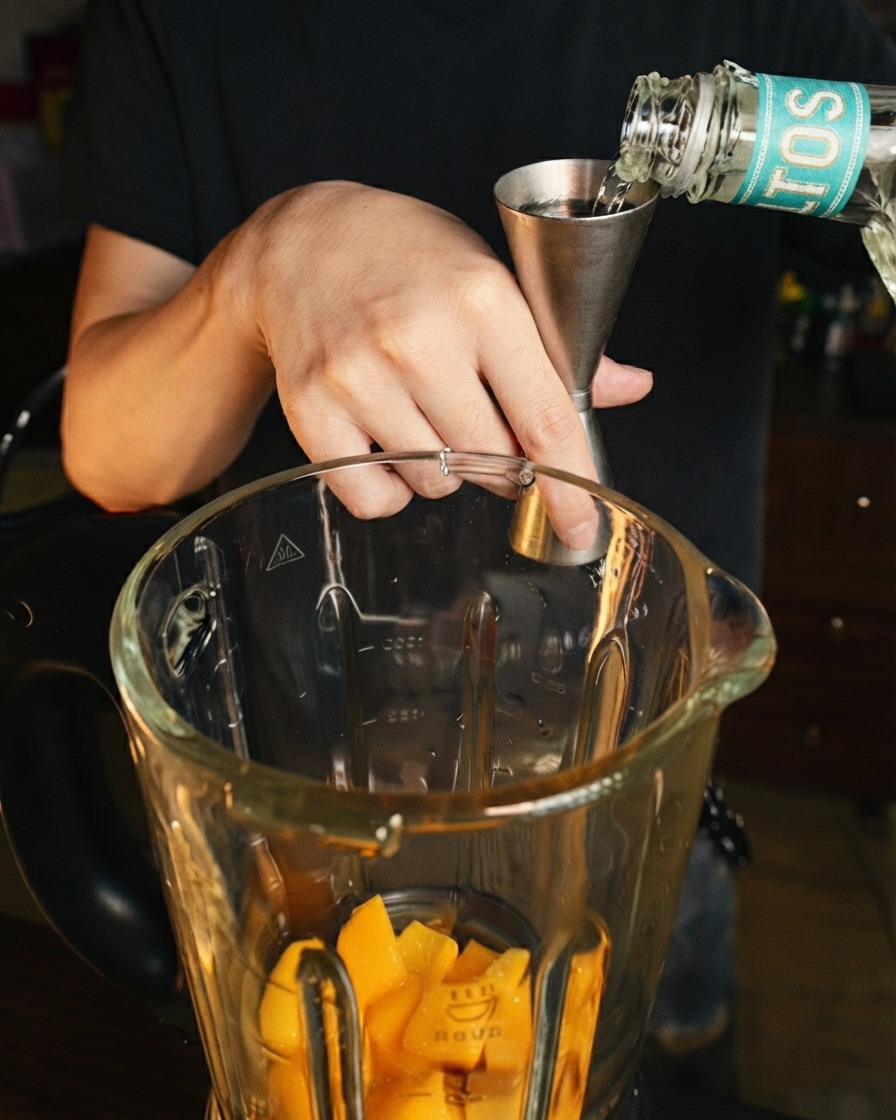 Liquore chiaro versato da un misurino in acciaio inossidabile in un boccale di frullatore sopra pezzi di mango fresco.