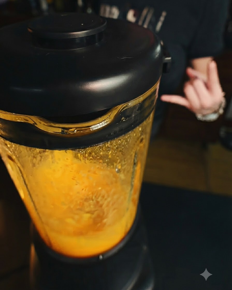 Un frullatore in funzione con un composto di mango arancione brillante allinterno, con una mano che fa un gesto sullo sfondo.