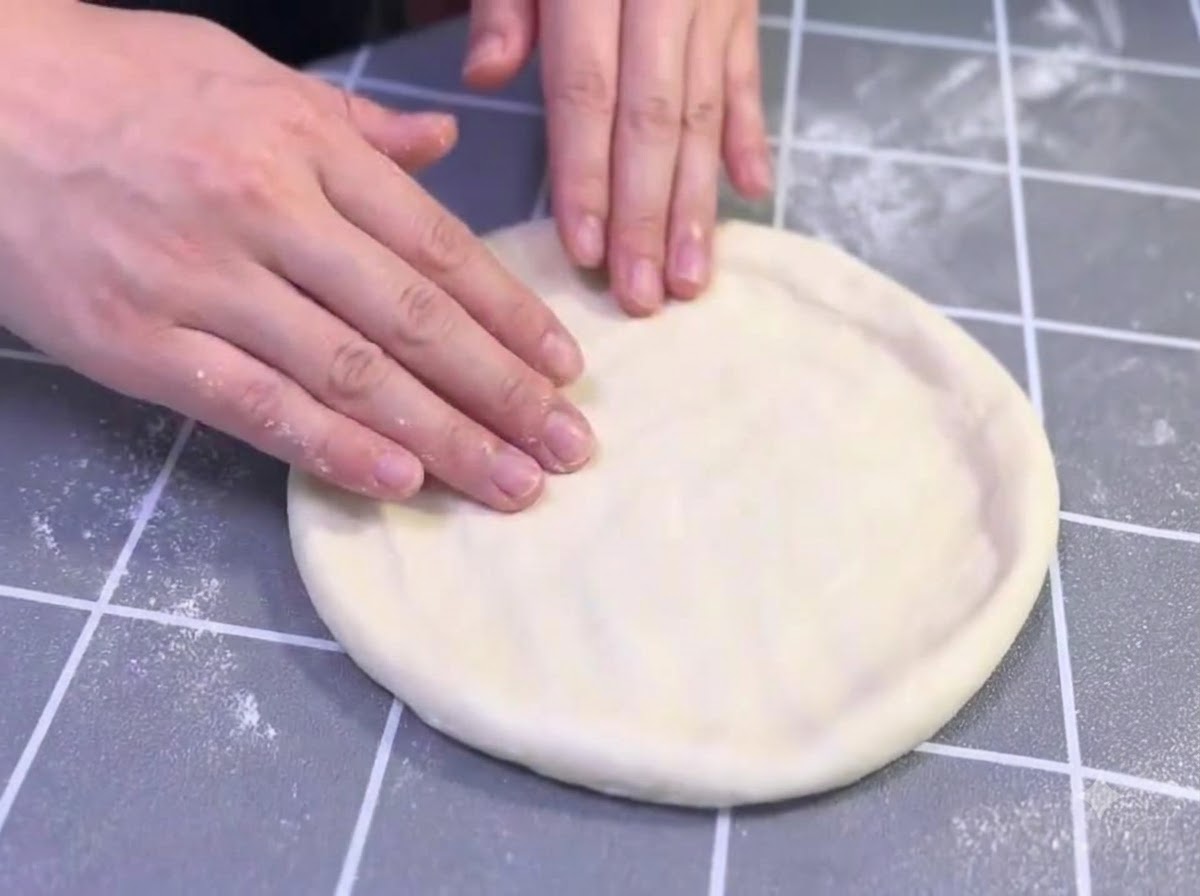 Mani che allungano e formano delicatamente un disco di pasta per pizza oliato su una superficie grigia a griglia.