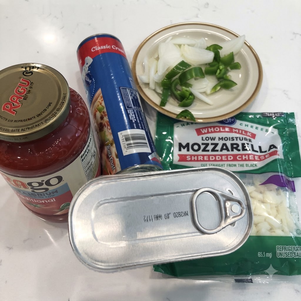 Una lattina di acciughe, un barattolo di salsa marinara, impasto per pizza in scatola, mozzarella grattugiata e cipolle e peperoni affettati su un piano di lavoro bianco.
