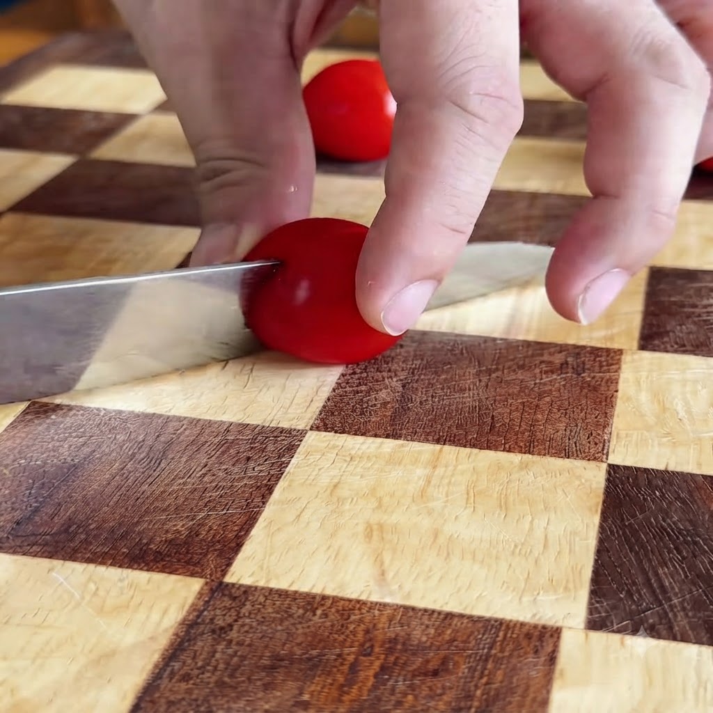 Mani che tagliano a metà un pomodorino rosso con un coltello su un tagliere a quadretti.