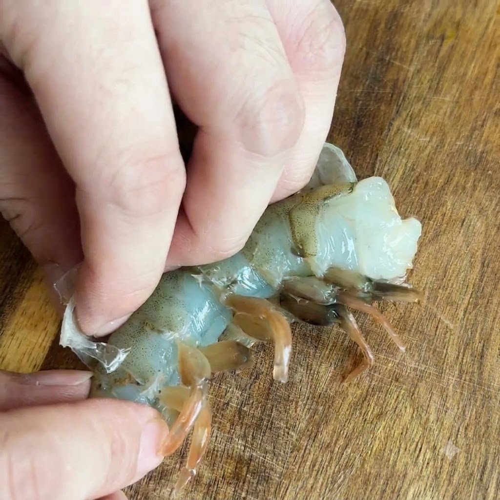 Dita che tirano via il guscio traslucido da un gambero crudo su una superficie di legno.