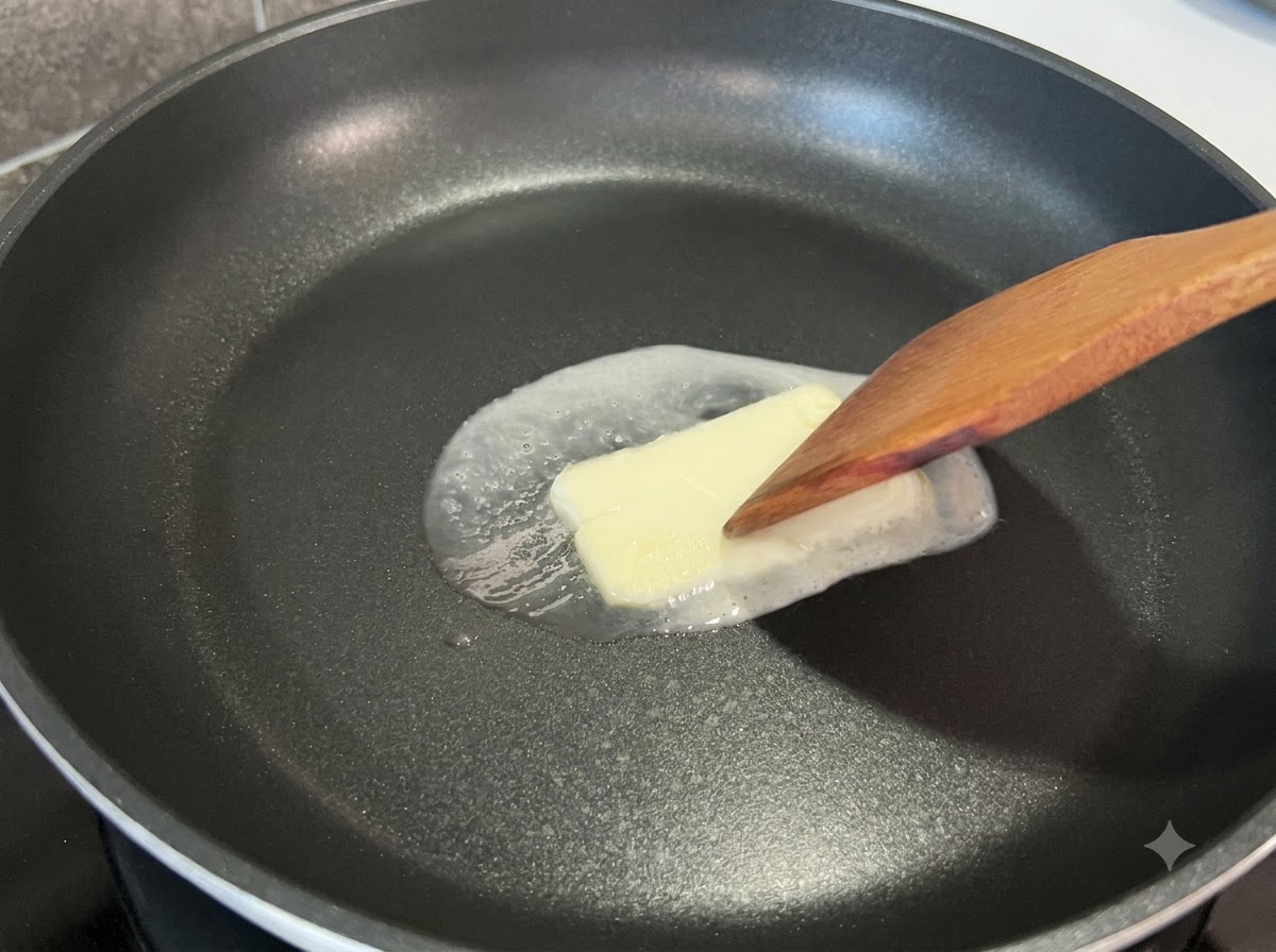 Una spatola di legno che preme leggermente su un panetto quadrato di burro che si scioglie in una padella nera.