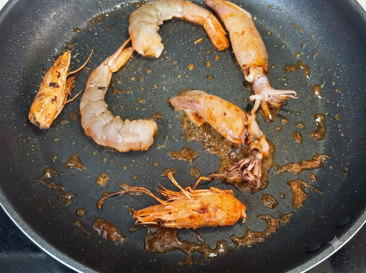 Gamberi, teste di gamberi intere e pezzi di calamaro che friggono in padella, conditi con macchie scure di pepe nero.
