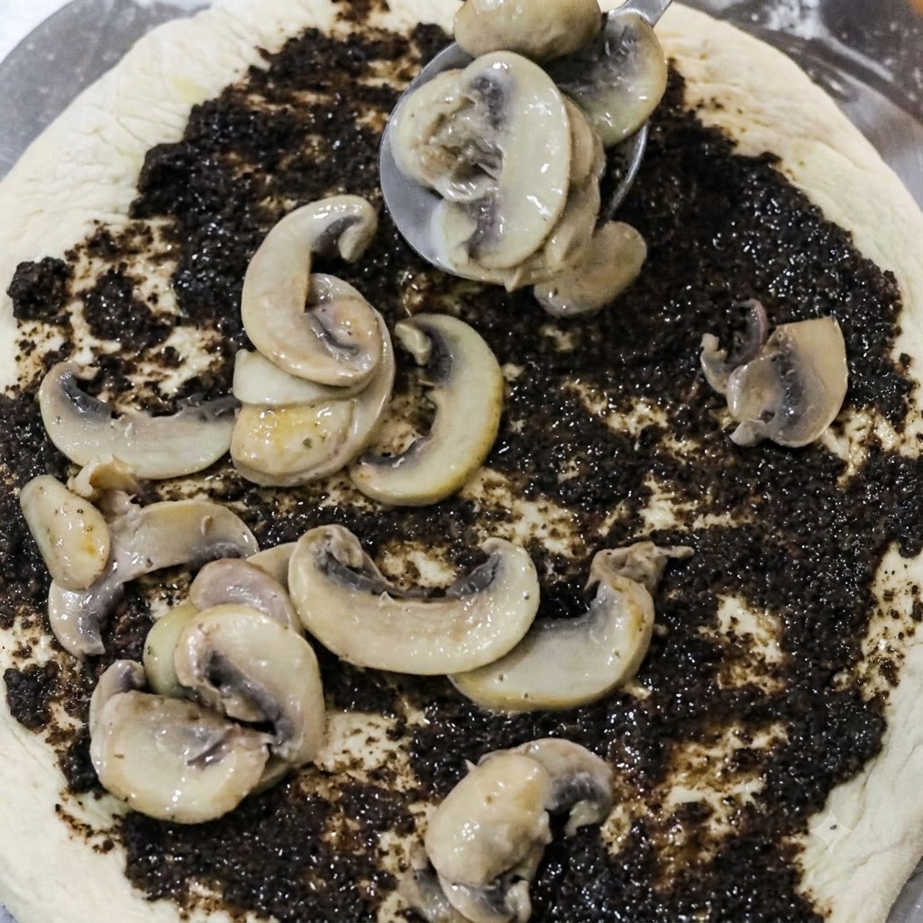 Cucchiaio che posiziona fette di funghi cotti su una base di pizza ricoperta di crema di tartufo nero.