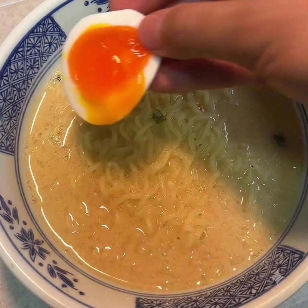 Una mano tiene metà di un uovo alla coque sopra una ciotola di noodles.
