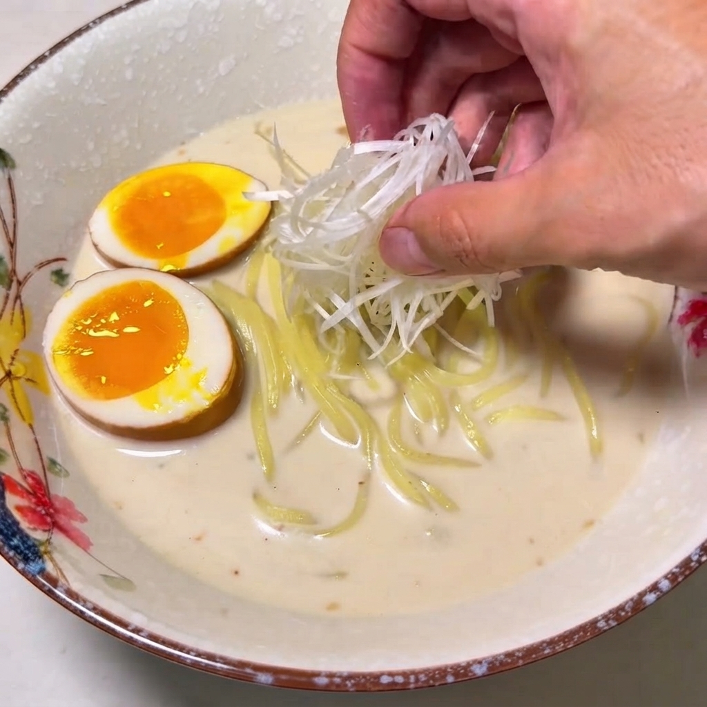 Una mano che posiziona un mucchietto di cipollotti bianchi sminuzzati su una ciotola di ramen.