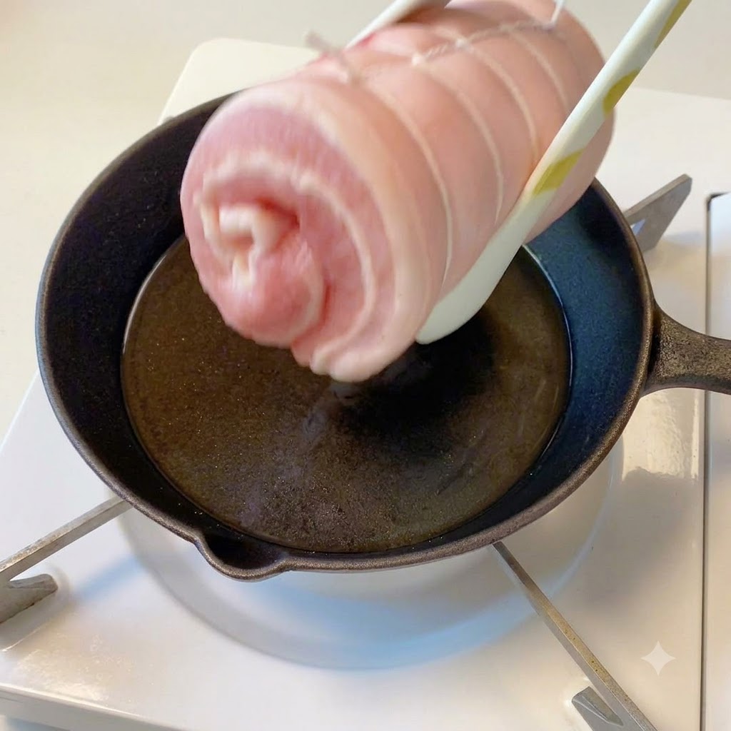 Il cilindro di pancetta di maiale legato tenuto con le pinze sopra una padella calda con olio.