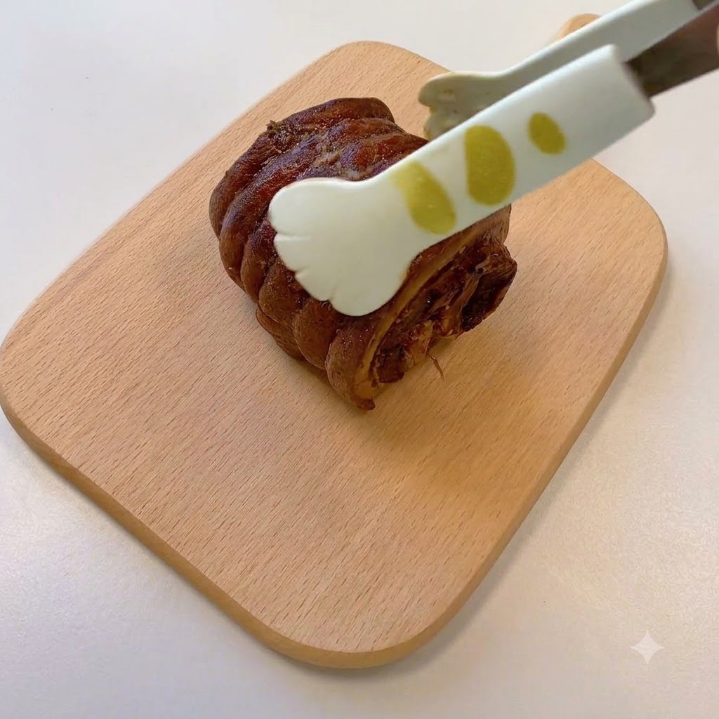 Un paio di pinze che tengono un pezzo di maiale chashu cotto e arrotolato adagiato su un tagliere di legno.