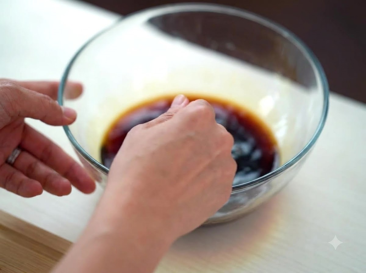 Una mano che mescola una salsa scura a base di soia in una ciotola di vetro trasparente.
