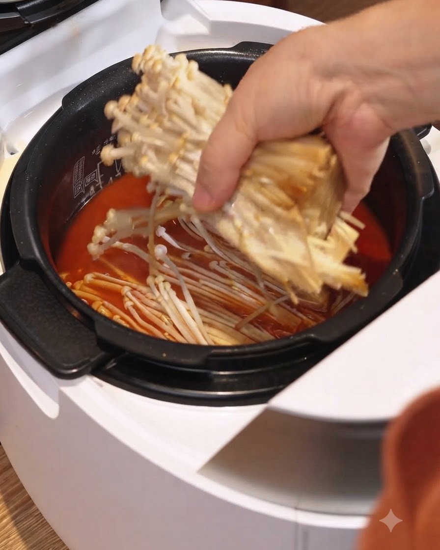 Un mazzetto di funghi enoki posizionato nel brodo rosso dello stufato allinterno di un cuociriso elettrico bianco.