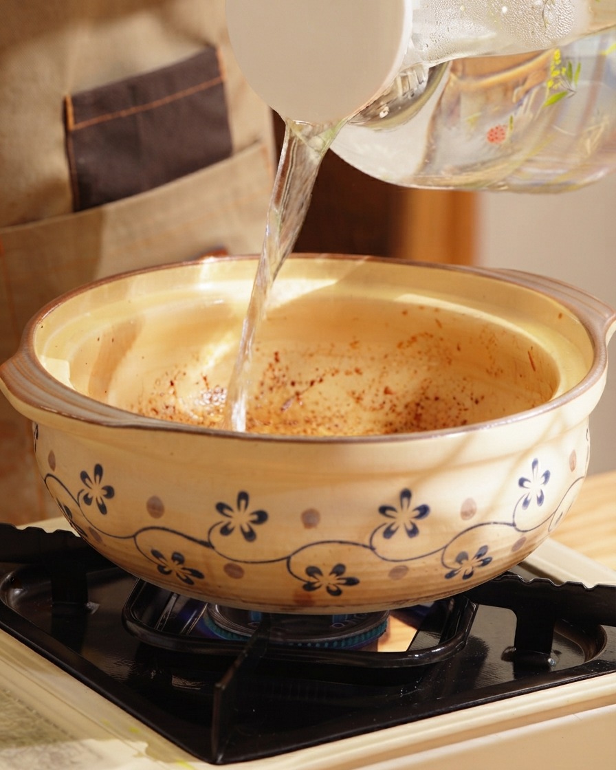 Acqua calda limpida versata da una brocca in una pentola di terracotta decorata su un piano cottura.