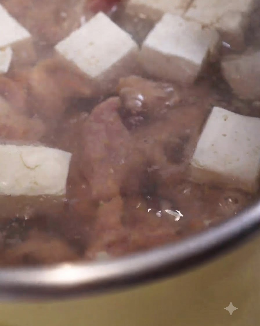 Fette di maiale marinato che cuociono in una pentola di zuppa che bolle accanto a cubetti di tofu bianco.