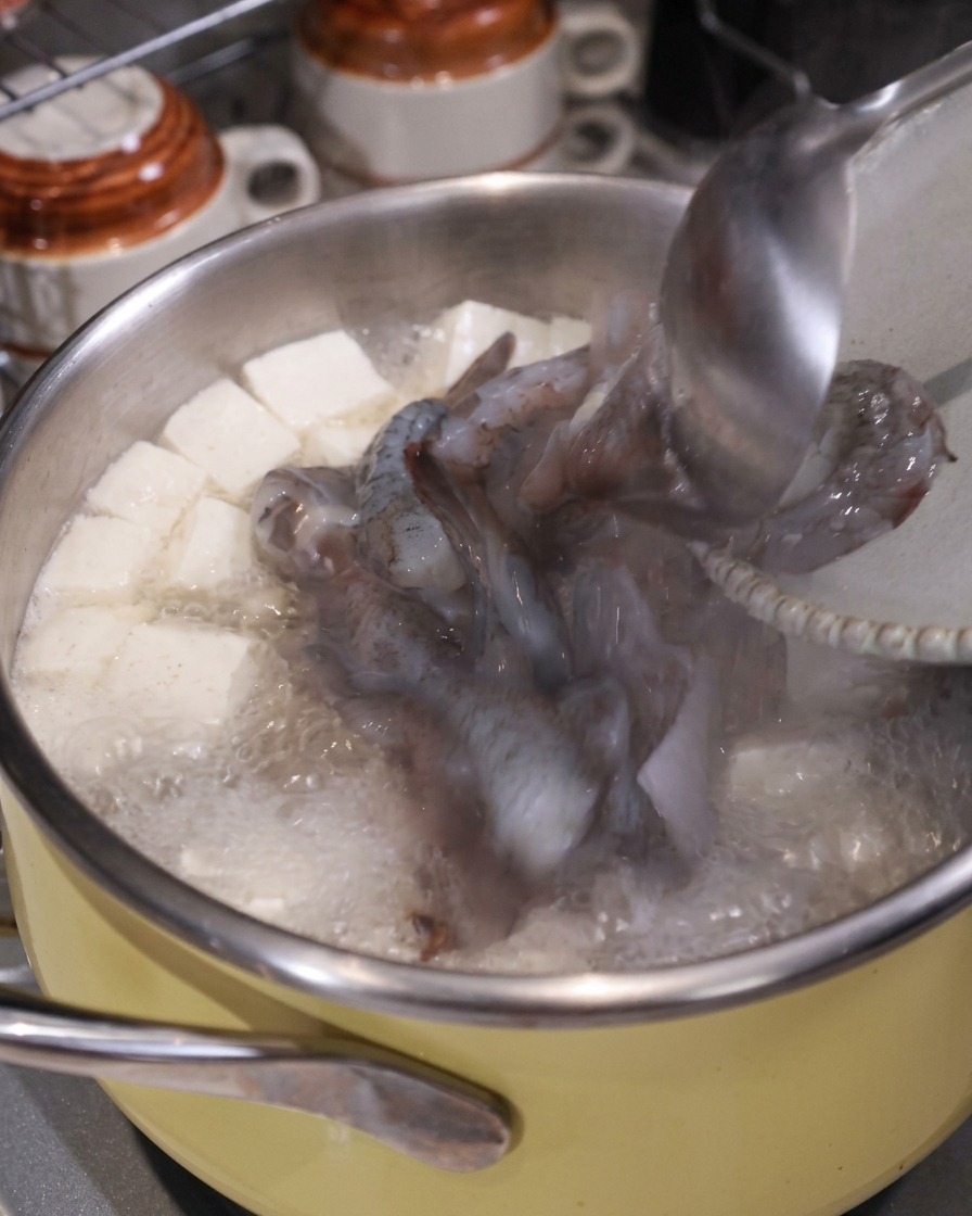 Un mestolo che aggiunge pezzi di calamaro crudo e gamberi in una pentola bollente di zuppa con tofu e maiale.