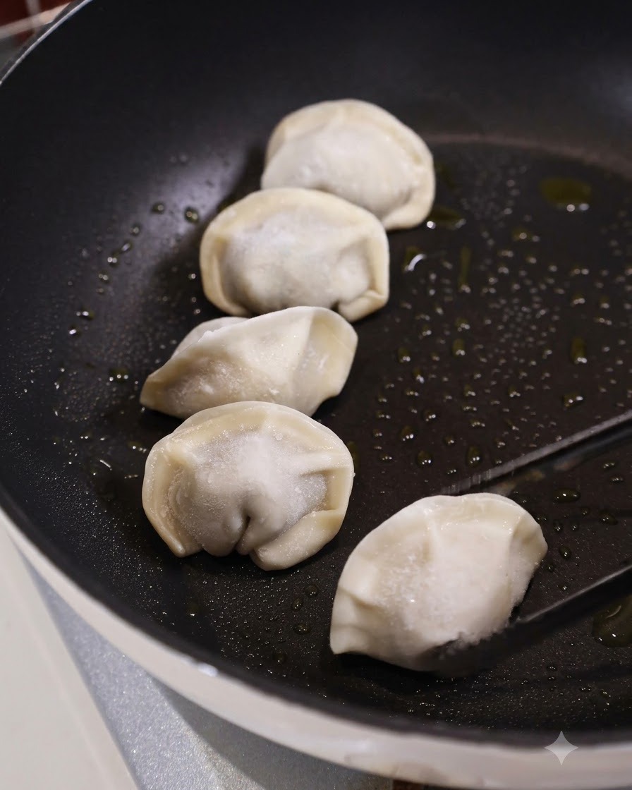 Le pinze da cucina posizionano un raviolo surgelato crudo in una padella piatta scura e oliata accanto a diversi altri ravioli.