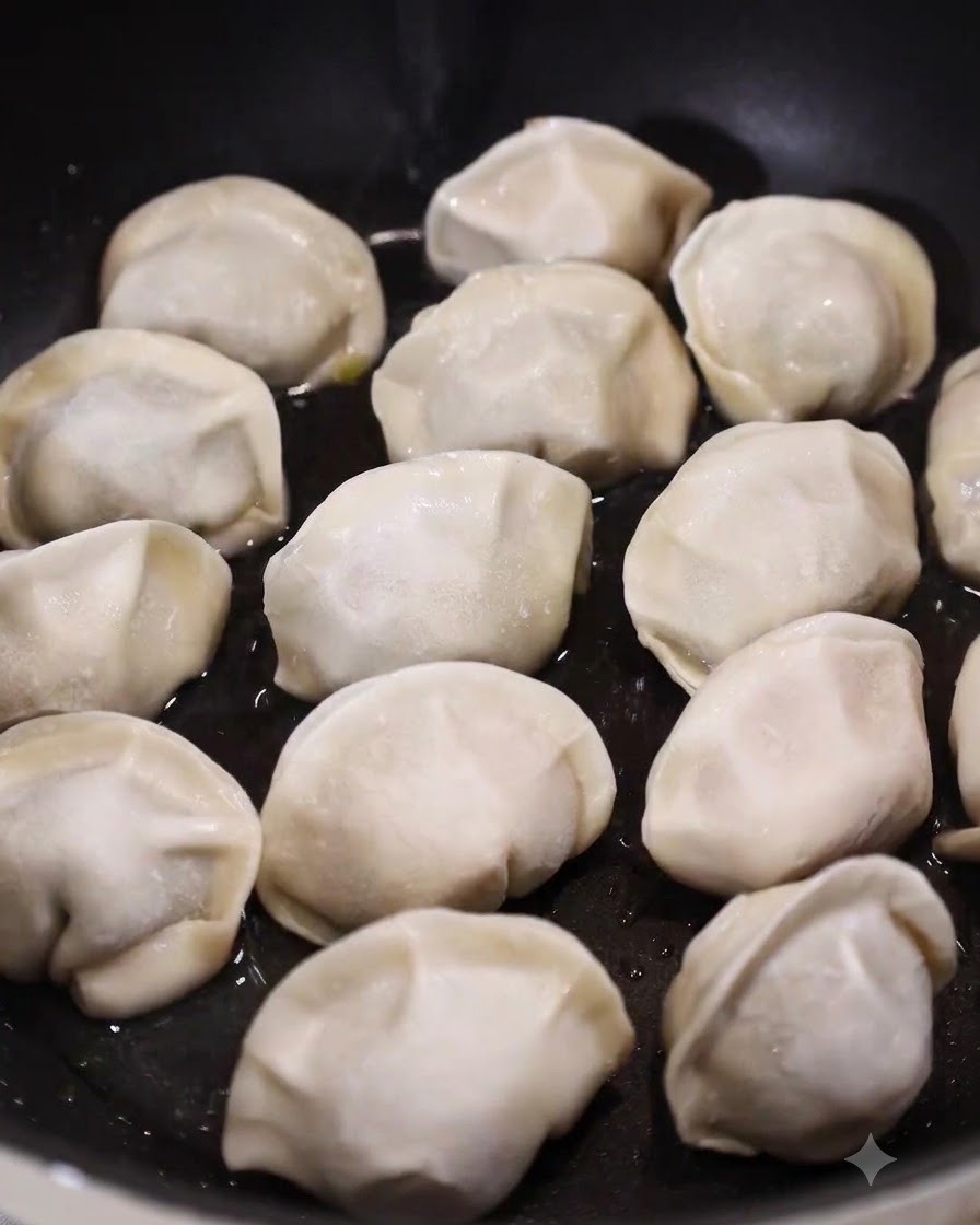 Una padella piatta piena di ravioli crudi adagiati in un sottile strato di acqua bollente e olio.