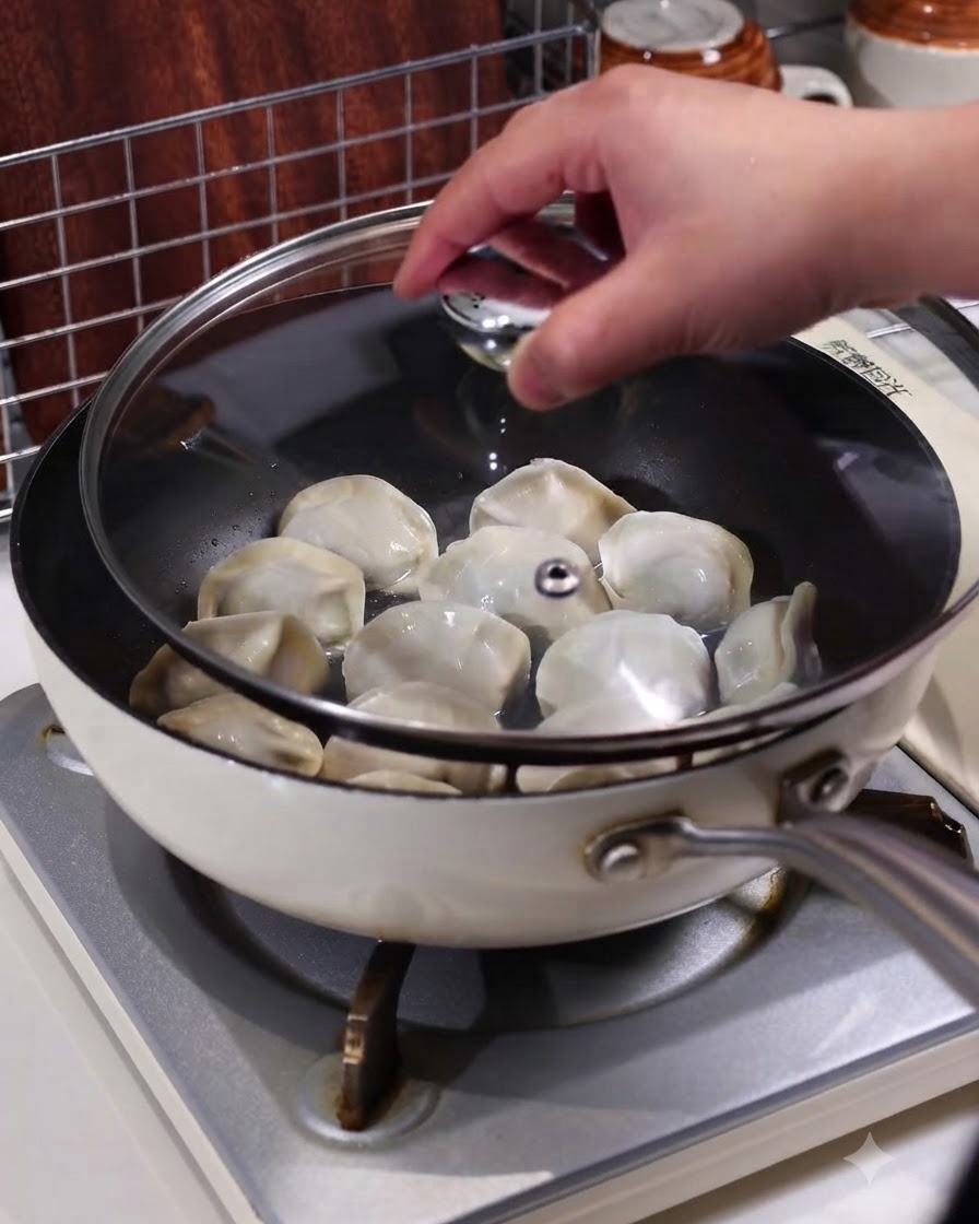 Una mano che posiziona un coperchio di vetro trasparente su una padella contenente ravioli fumanti su un piano cottura.