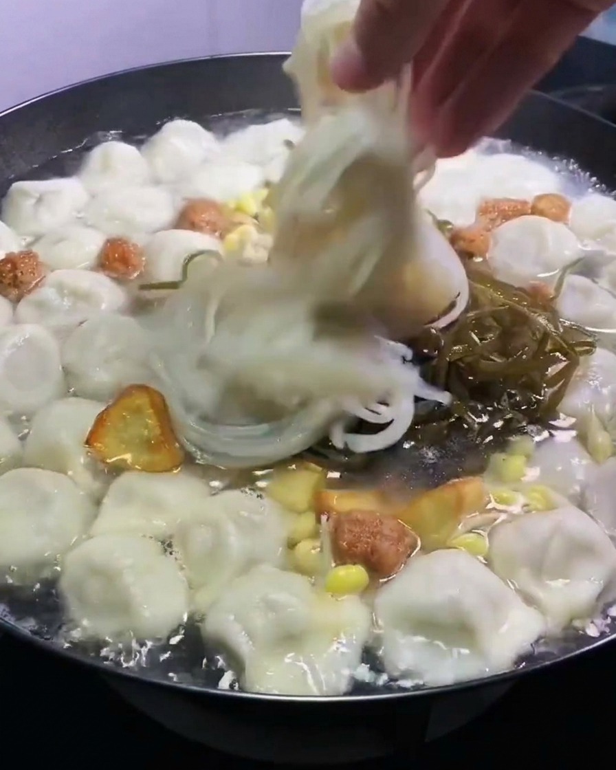 Una mano che posiziona alga kelp sminuzzata e noodles di vetro traslucidi in una pentola di dumpling bollenti e brodo.