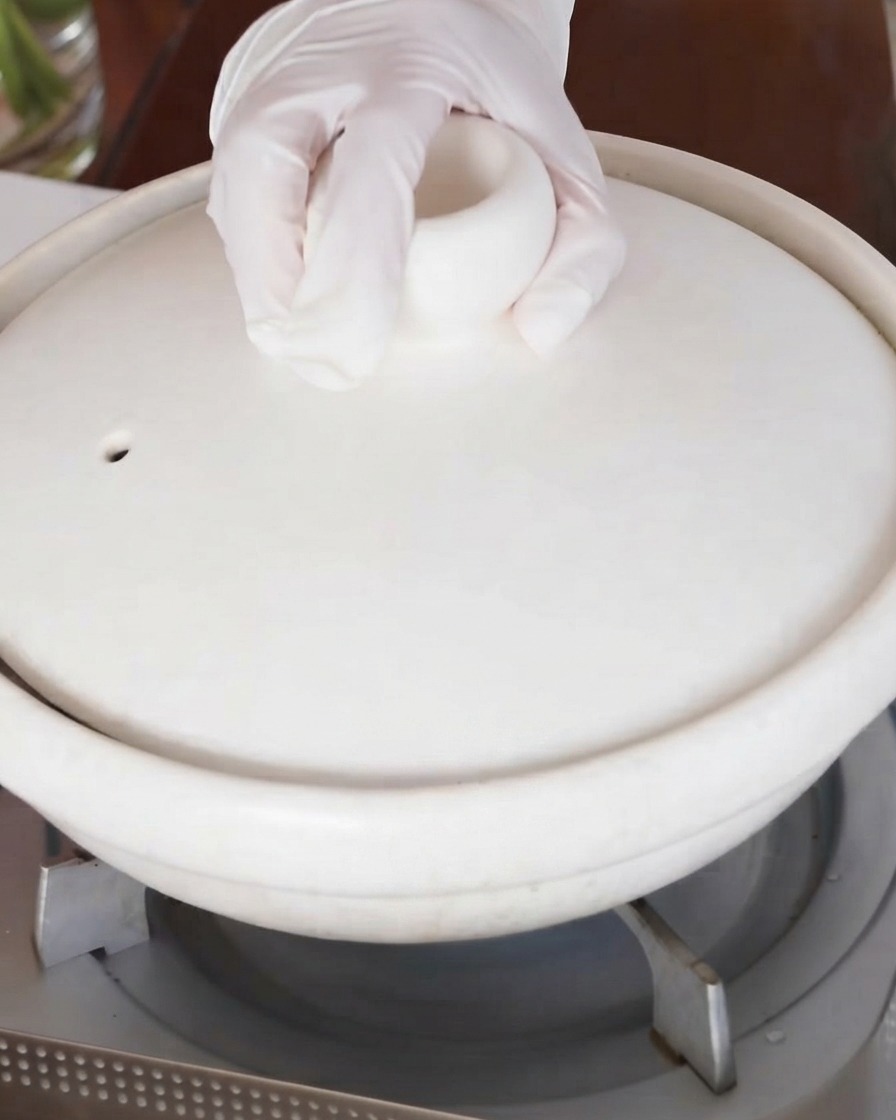 Una mano che posiziona il coperchio sulla pentola di brasatura in ceramica bianca sul fuoco.