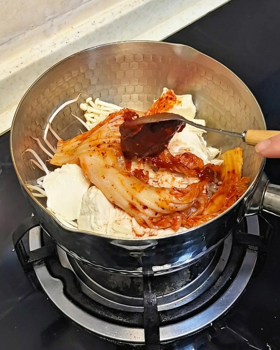 Un cucchiaio che aggiunge una dose di pasta di peperoncino coreana rossa densa sugli ingredienti nella pentola.