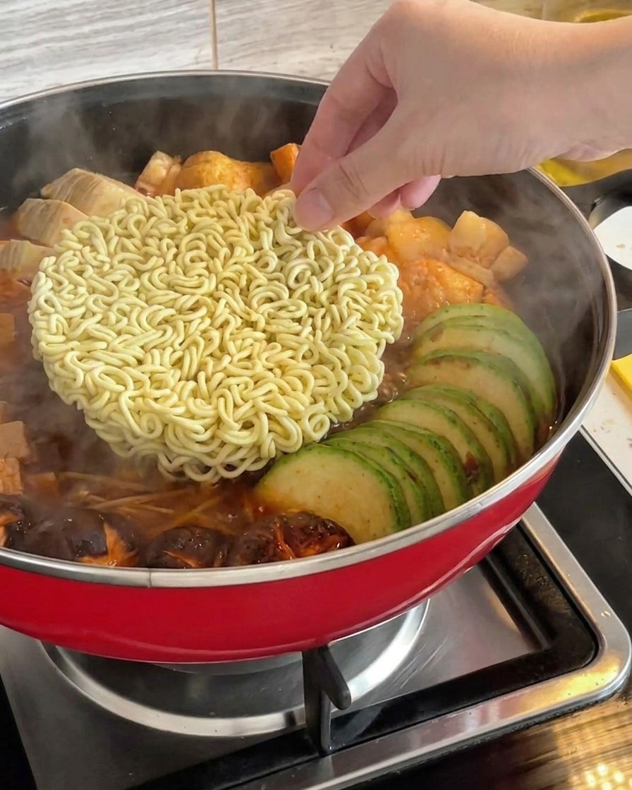 Una mano posiziona un panetto di ramen istantaneo secco al centro dello stufato coreano in ebollizione.
