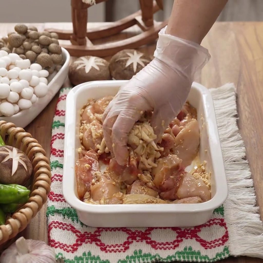 Una mano guantata che mescola e massaggia la marinata nei pezzi di pollo allinterno di un piatto bianco.