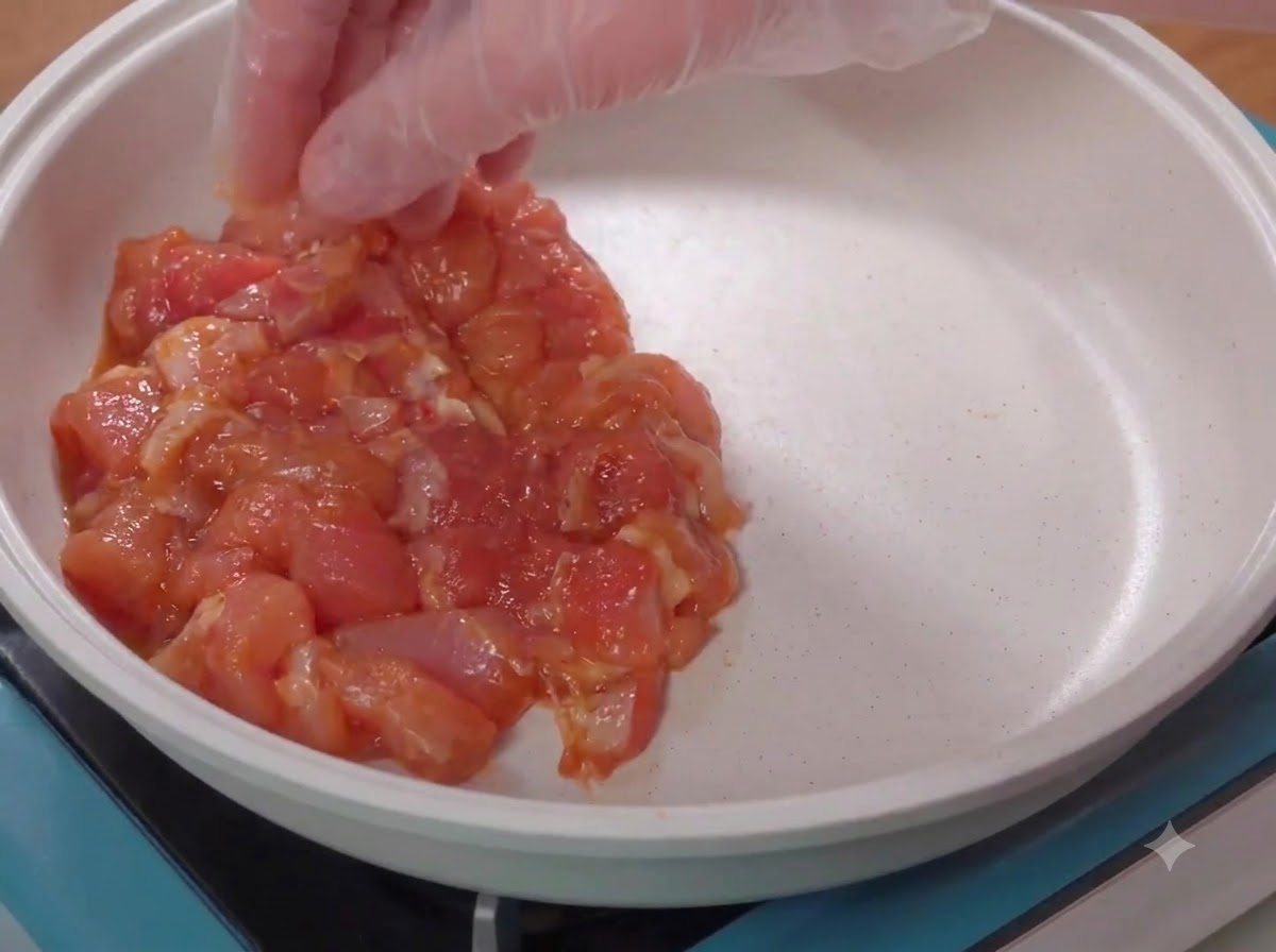 Una mano con guanto che dispone pezzi di pollo crudo marinato in una padella bianca.