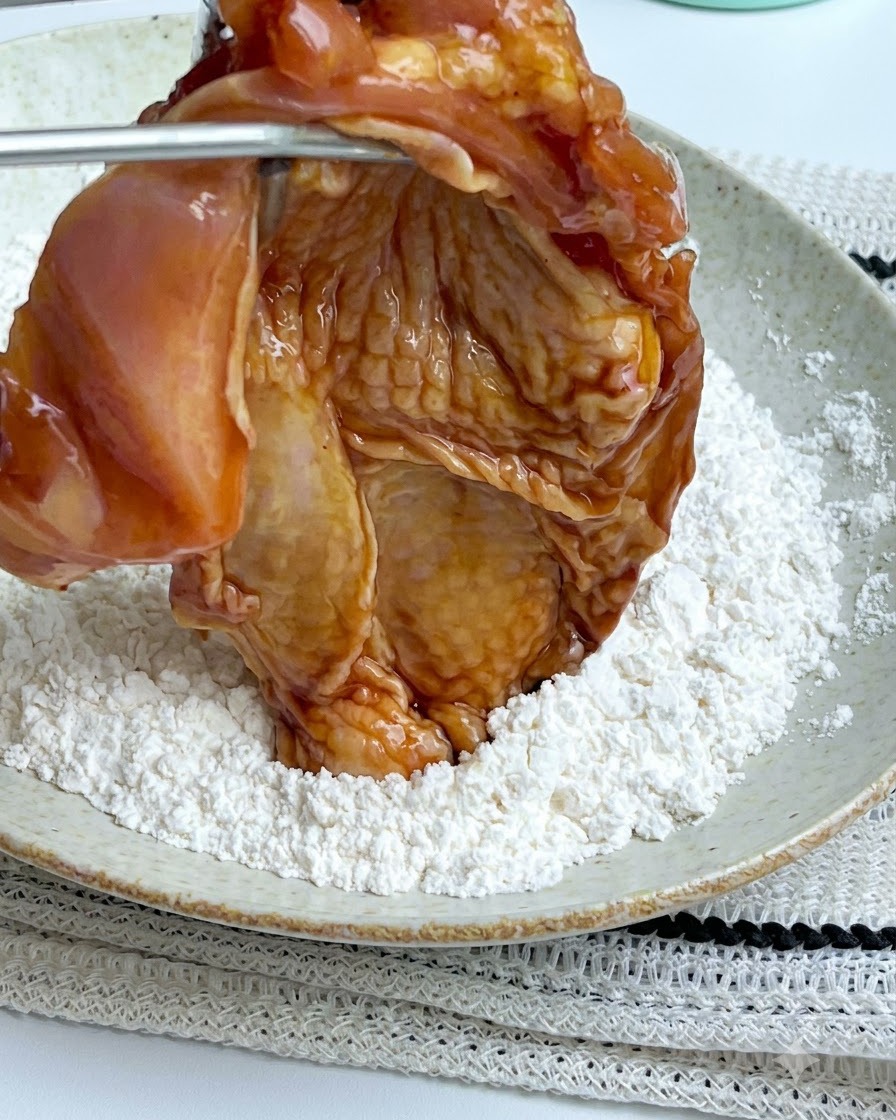 Pinze che tengono un pezzo di pollo marinato sopra un piatto bianco pieno di amido di mais in polvere.