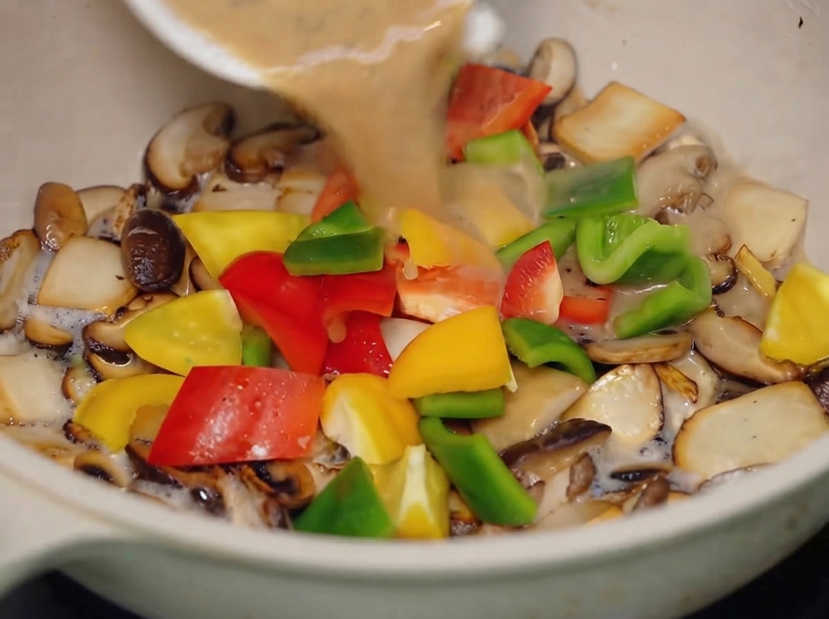 La salsa al pepe nero versata da una piccola ciotola bianca nella padella con funghi misti e peperoni colorati.