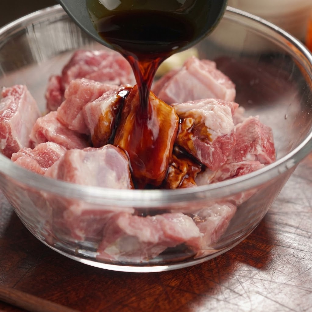 Salsa di soia scura versata da un mestolo su pezzi di costine di maiale crudi in una ciotola di vetro.