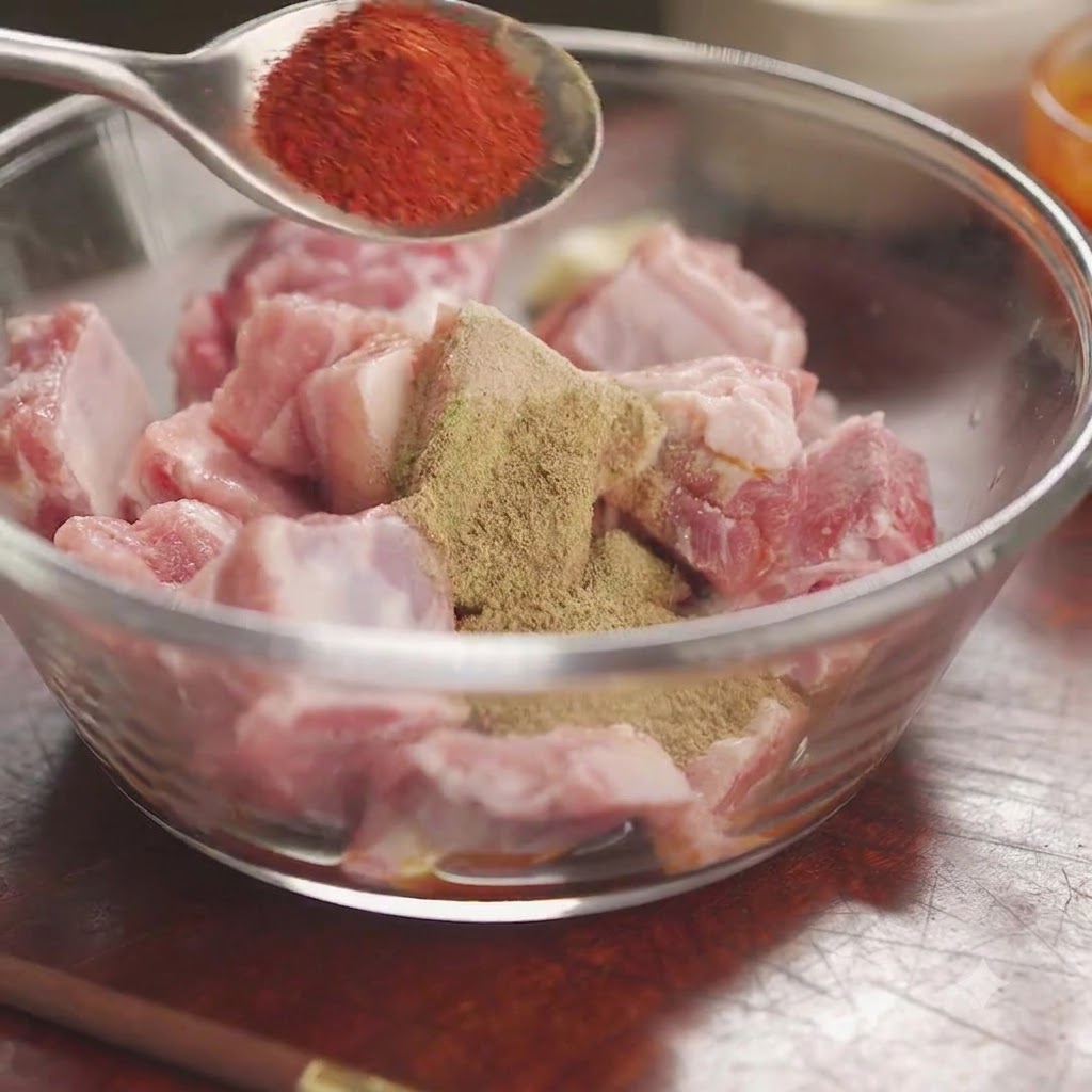Peperoncino rosso in polvere su un cucchiaio tenuto sopra una ciotola di costine di maiale crude coperte di spezie ed erbe miste.