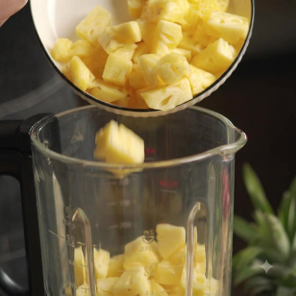 Pezzi di ananas fresco tagliati a cubetti versati da una ciotola bianca in un frullatore di vetro.