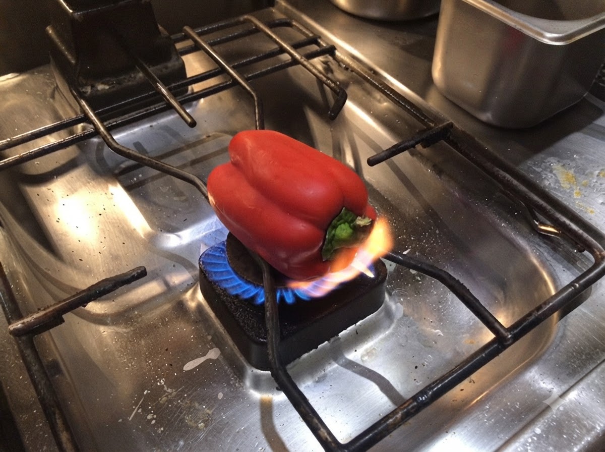 Un peperone rosso adagiato sul bruciatore di un piano cottura a gas con una fiamma viva che ne brucia la base.