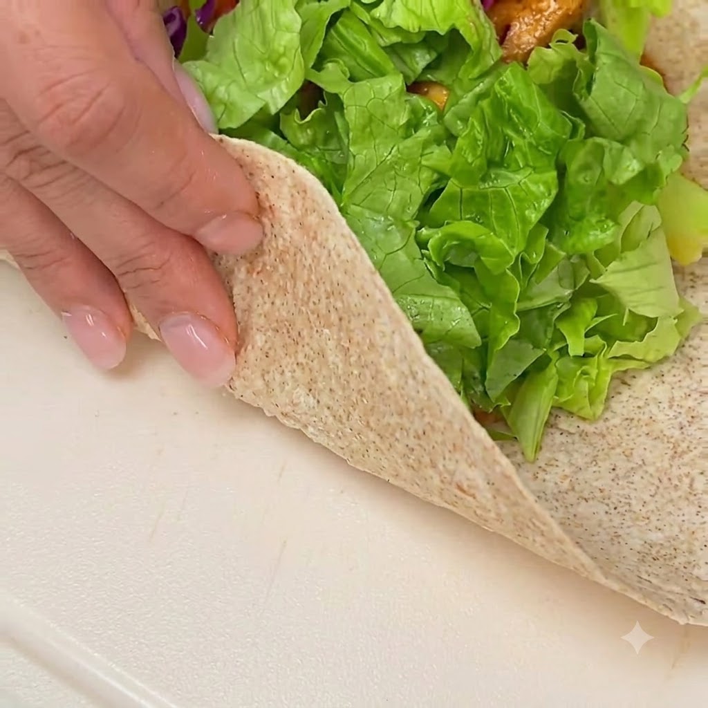 Mani che piegano e arrotolano una tortilla integrale farcita su una superficie bianca pulita.