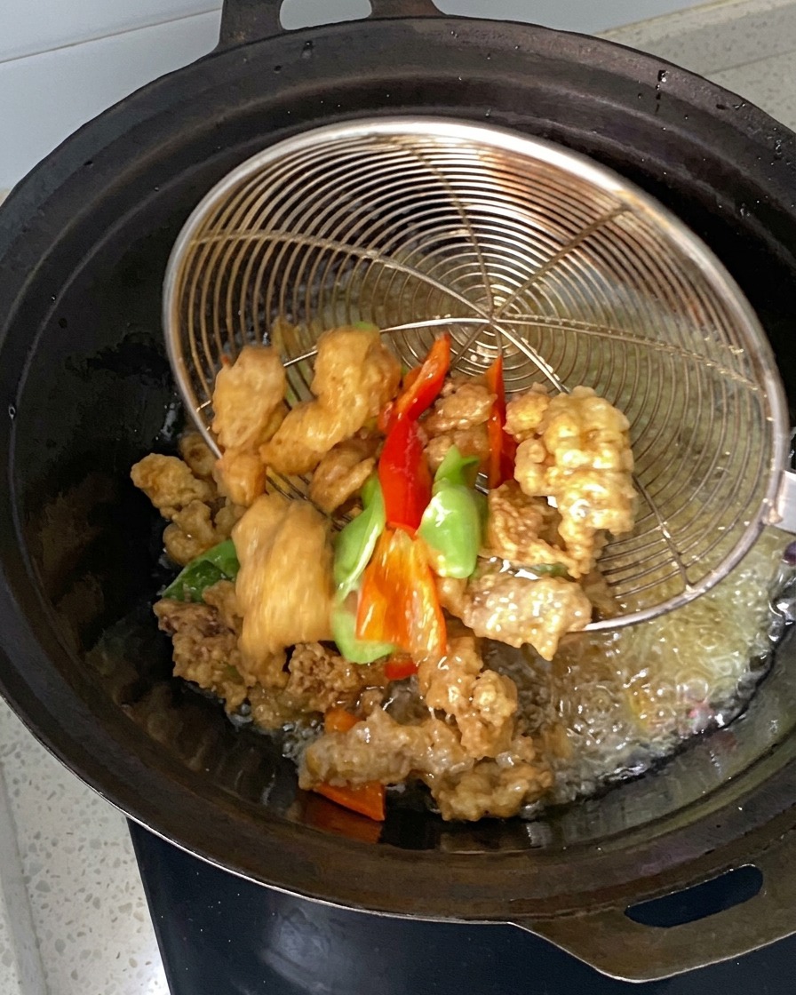 Pezzi di carne marinati e peperoncini immersi e raccolti con un colino a rete in un wok pieno di olio caldo.