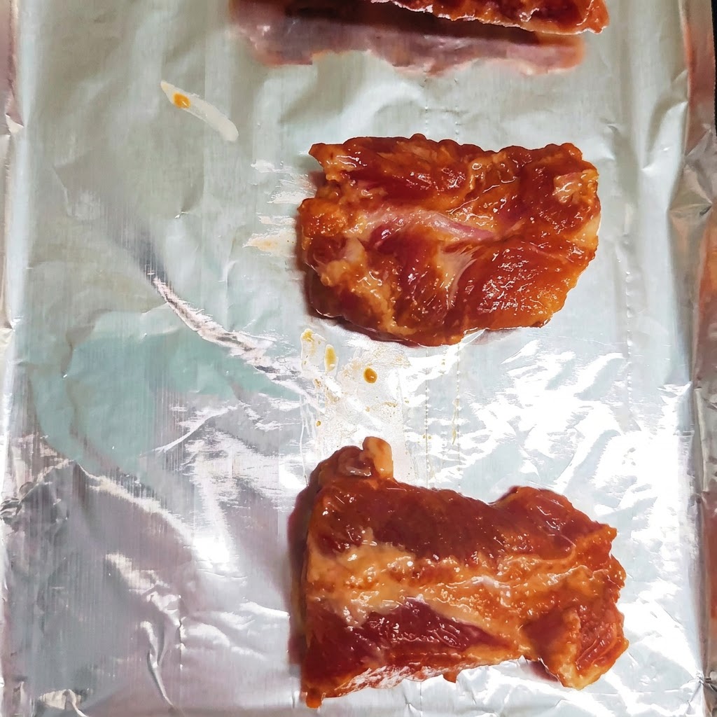 Pezzi di costine di maialeマリンate disposti ordinatamente in ununica fila su una teglia da forno foderata di alluminio.