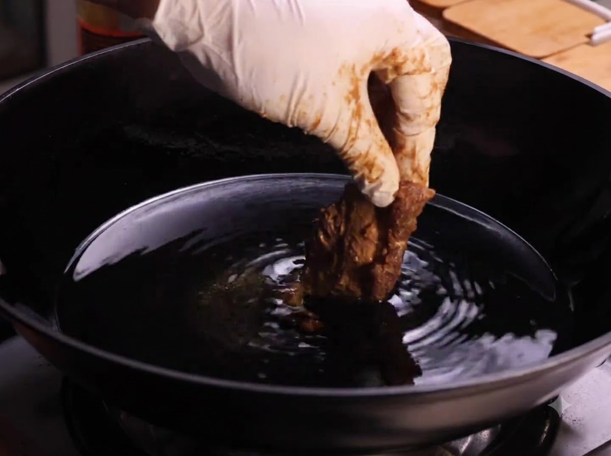 Una mano guantata che immerge un pezzo di costina di maiale infarinata in un wok pieno di olio caldo per friggere.