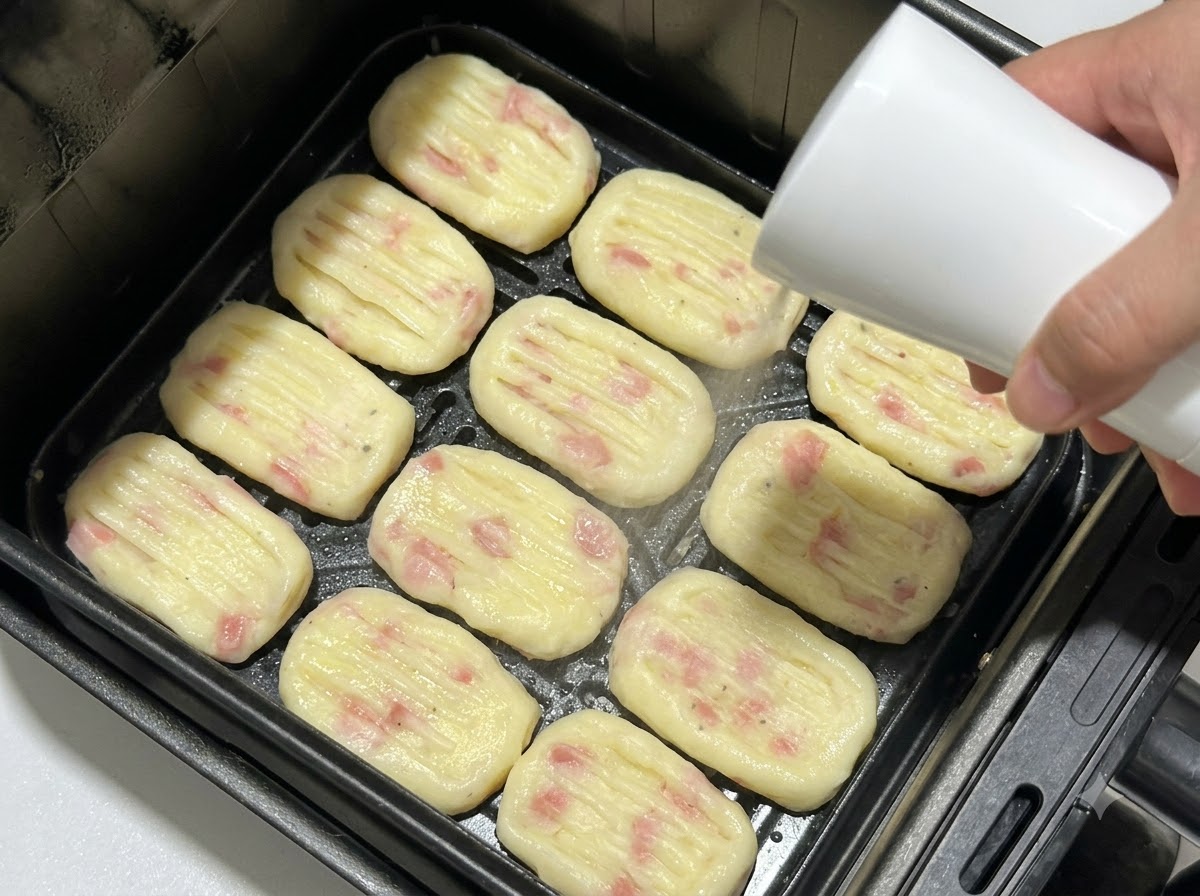 Crocchette di patate modellate disposte nel cestello di una friggitrice ad aria mentre vengono spruzzate con olio da un flacone spray.