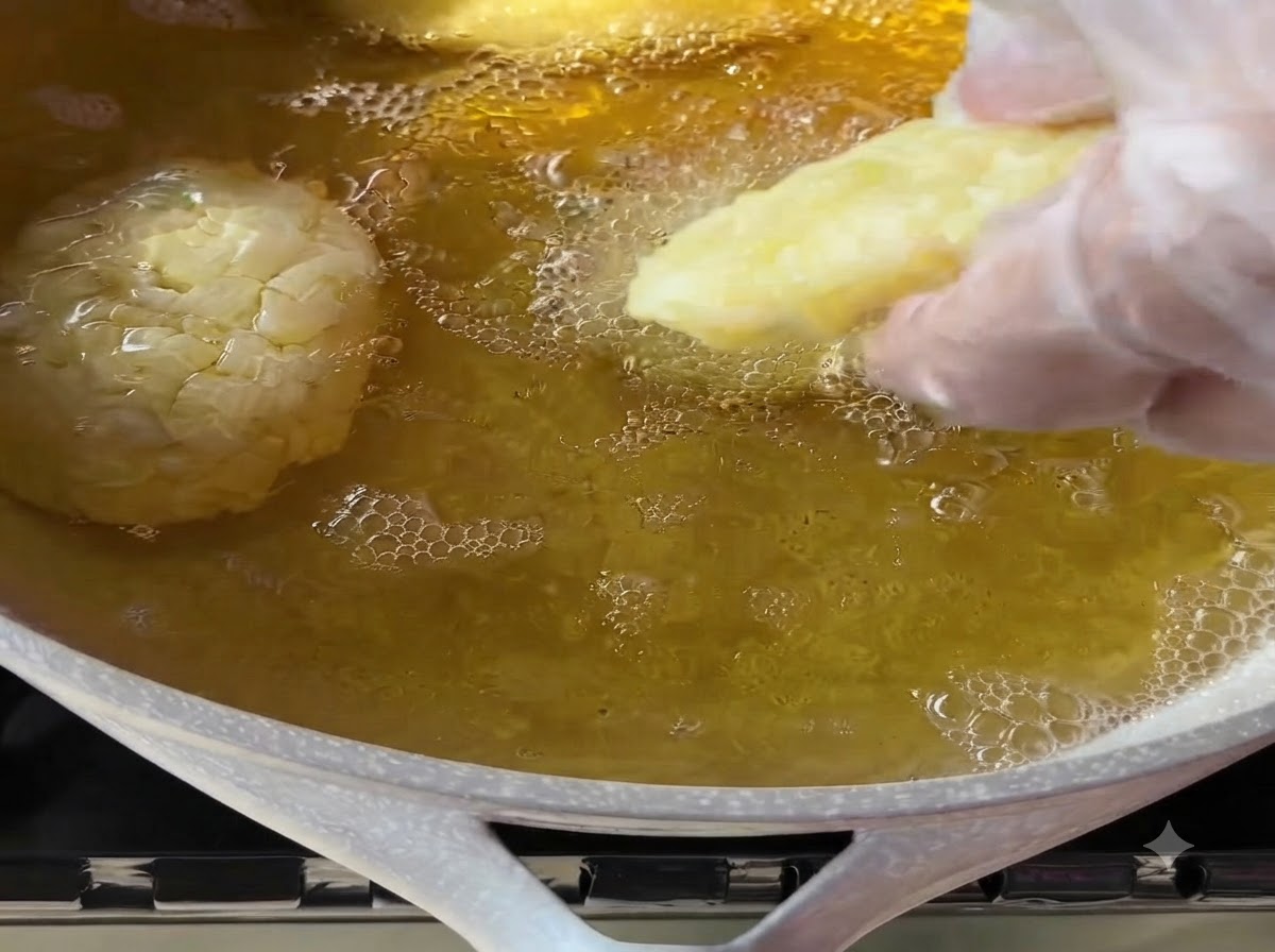 Una mano guantata che posiziona delicatamente una polpetta di patate modellata in una padella di olio da cucina caldo e bollente.