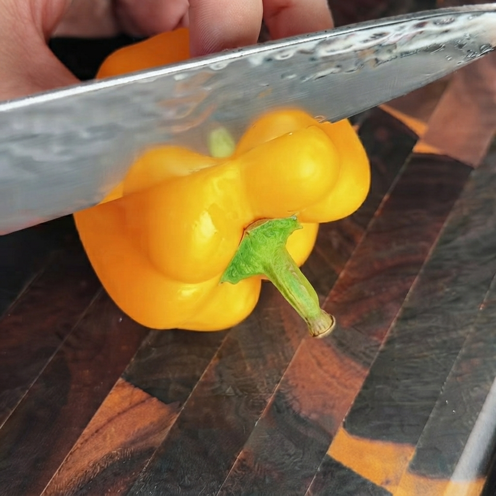 Un coltello affilato che taglia la sezione superiore di un peperone giallo brillante su un tagliere di legno.