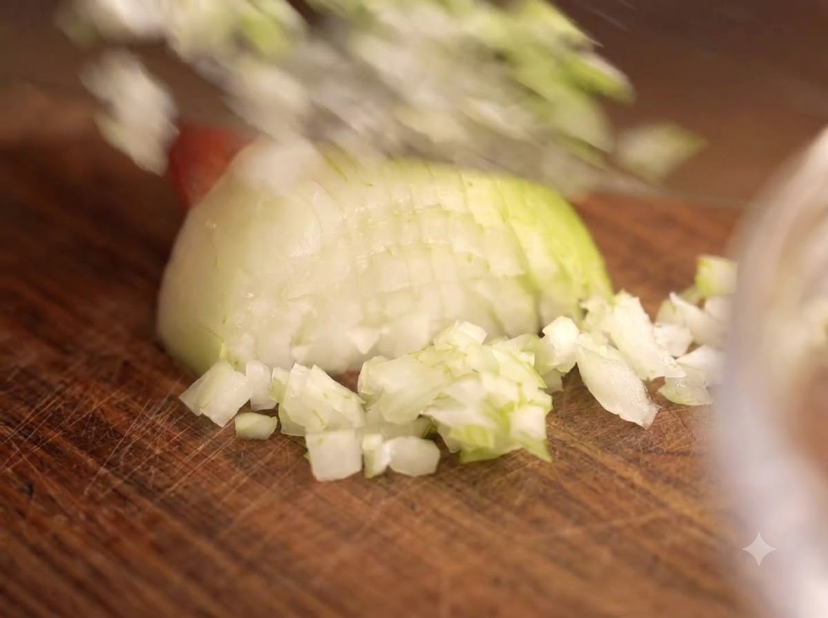 Un coltello che taglia cipolle e aglio su un tagliere di legno accanto a una ciotola di vetro di carne.
