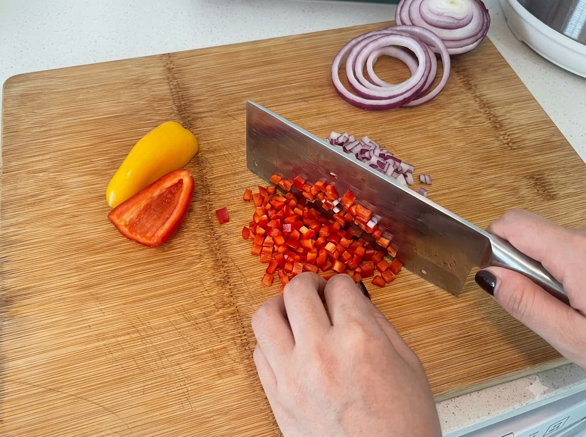 Peperoni rossi e gialli tagliati a cubetti su un tagliere di legno con una mannaia da cucina.