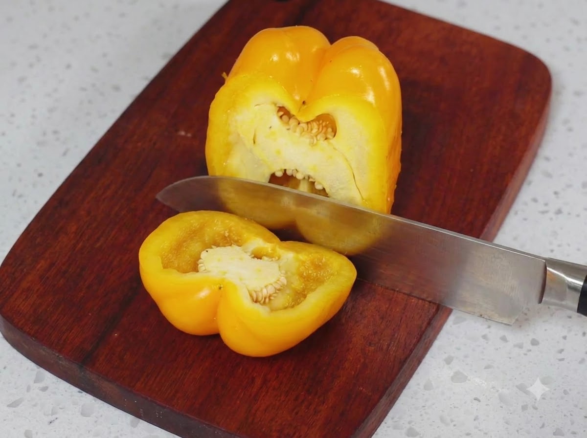 Un peperone giallo tagliato a metà su un tagliere di legno con un coltello da cucina.
