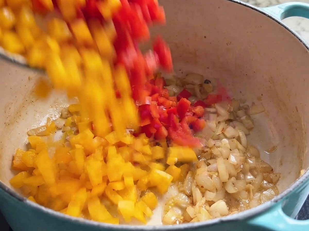 Cubetti di peperone rosso e giallo che cadono in una pentola di cottura piena di cipolle rosolate.